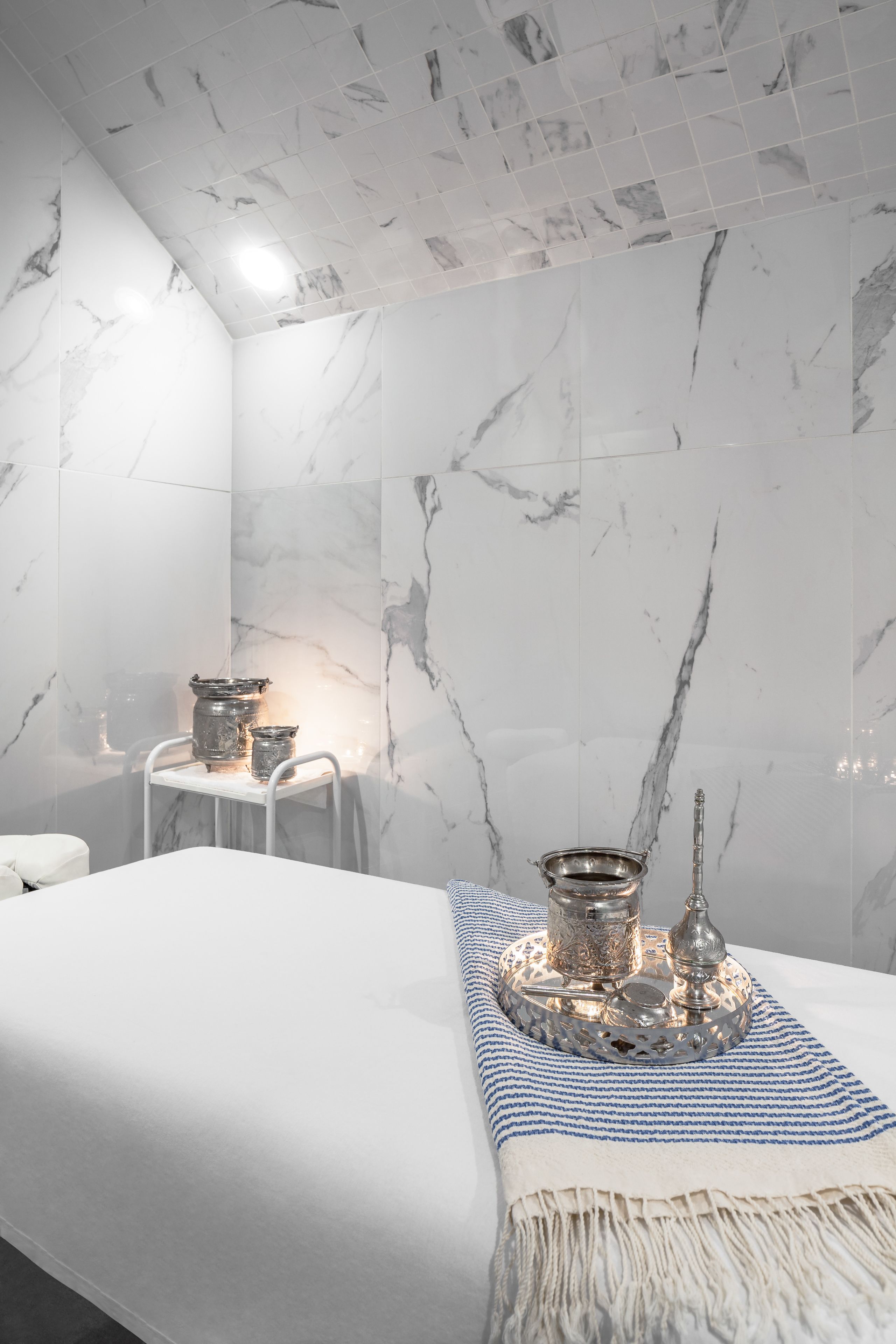 A hotel room with a marble wall, a bed with a white and blue striped blanket, and a small table with silver items.