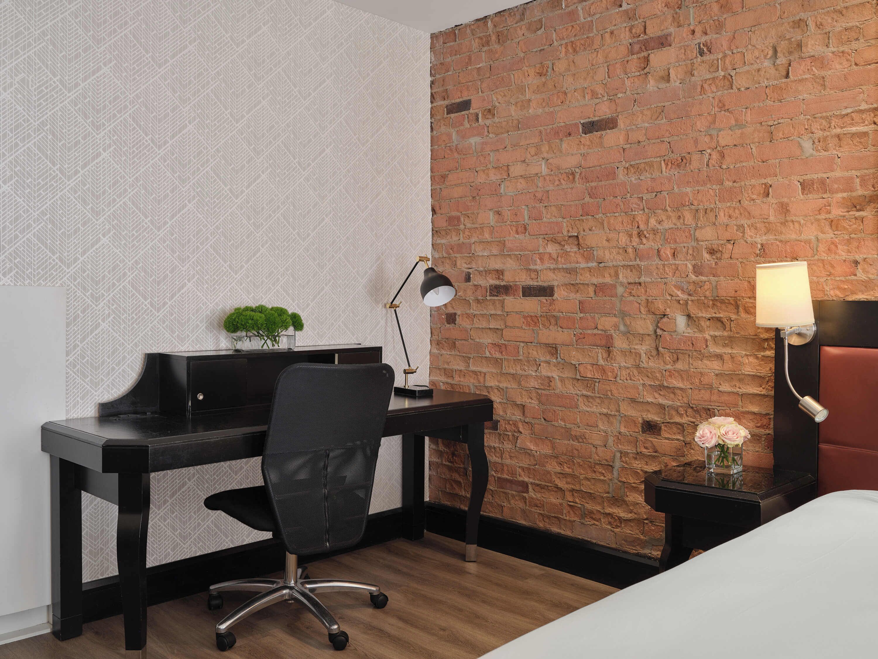 Una habitación de hotel con pared de ladrillo, un escritorio con computadora, una silla, una lámpara, una mesita con flores y una cama.