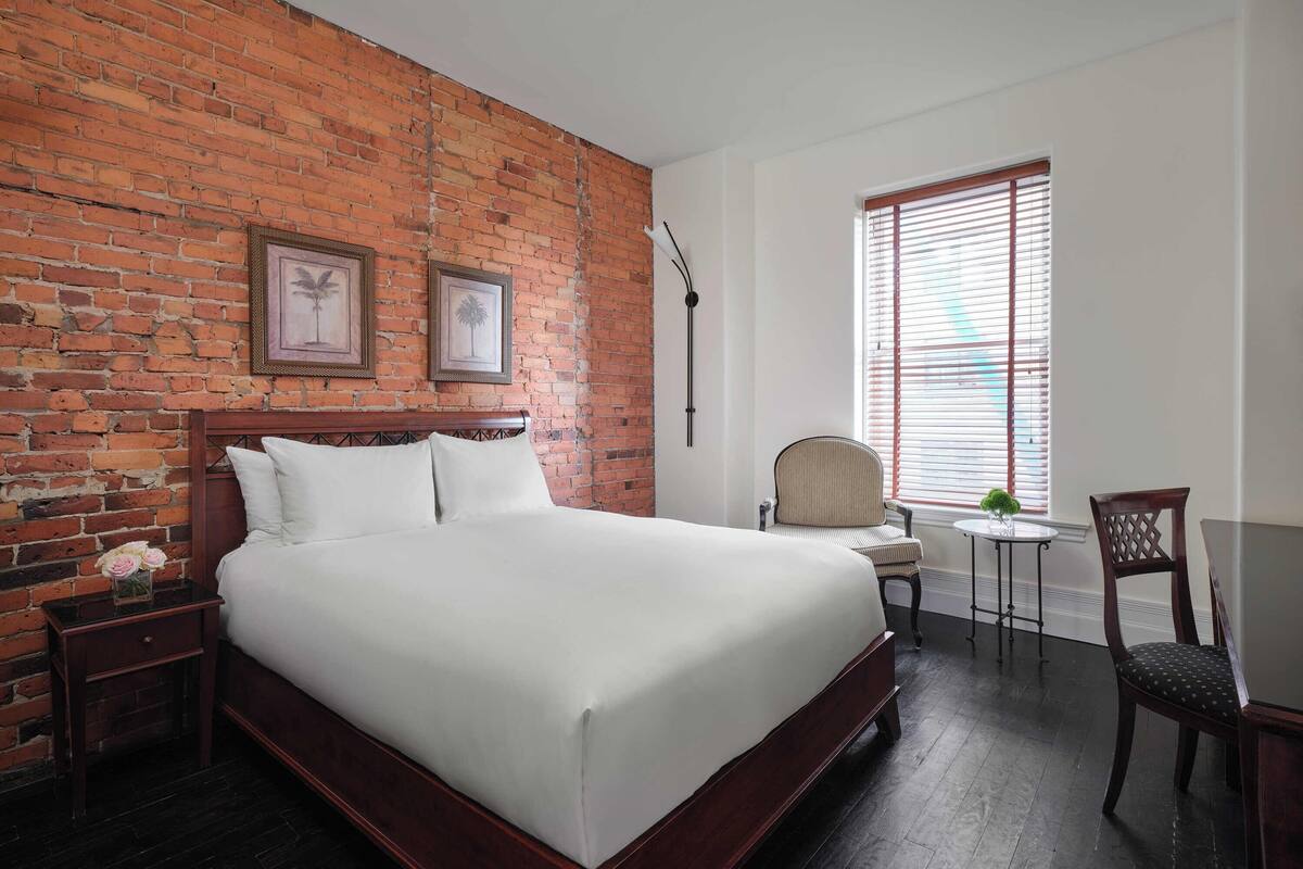 A bedroom with a brick wall, a bed, a chair, a small table, and a window with blinds.