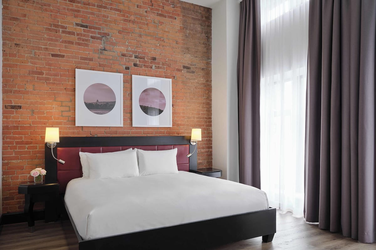 A hotel room with a brick wall, a bed with white bedding, two wall-mounted lamps, and a large window with curtains.