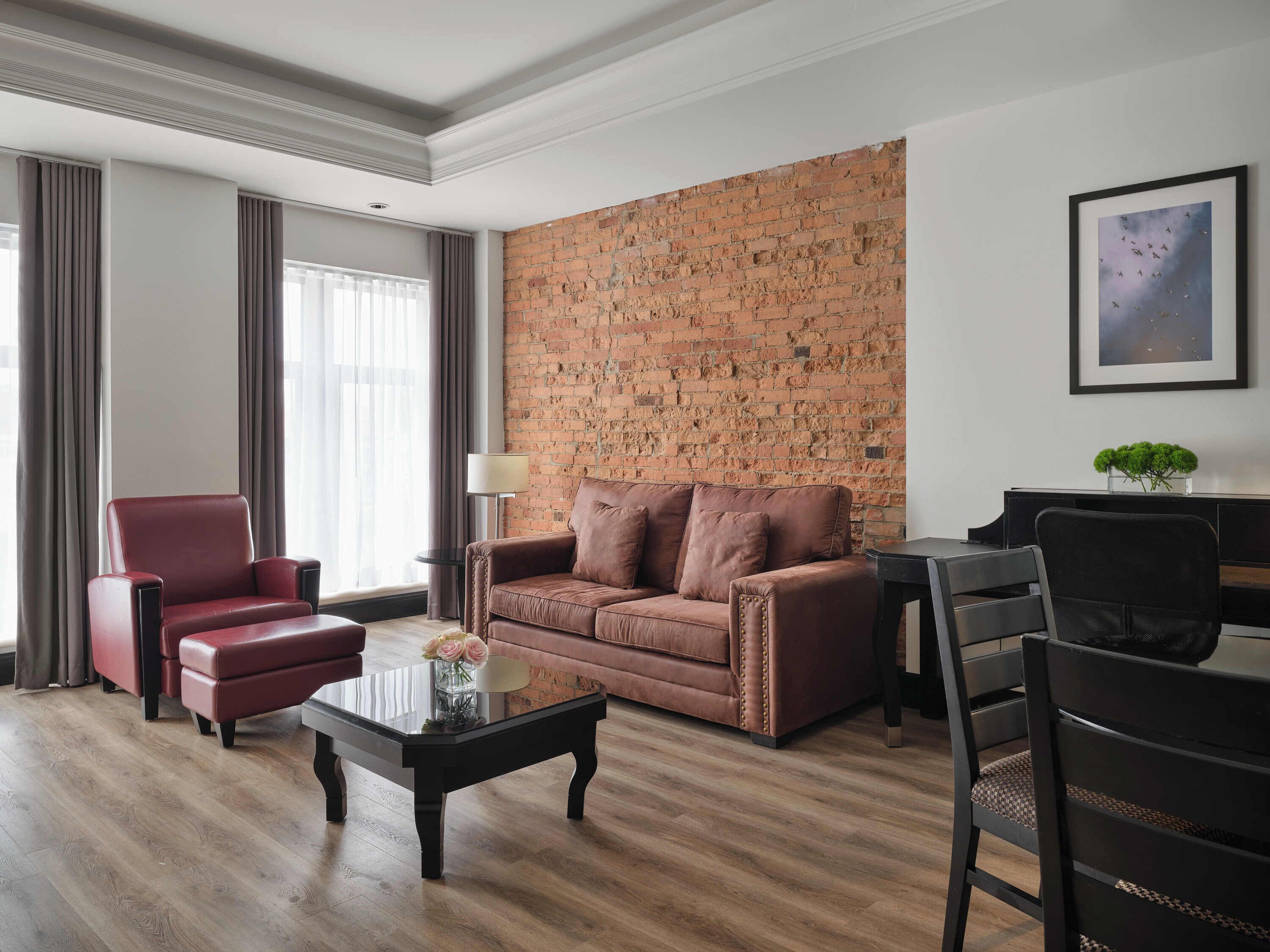 A modern living room with a brick accent wall, a leather sofa, a red armchair, a coffee table with flowers, and a piano.