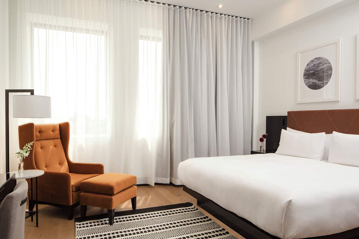A hotel room with a large bed, a chair, a small table with a lamp, and a framed picture on the wall.