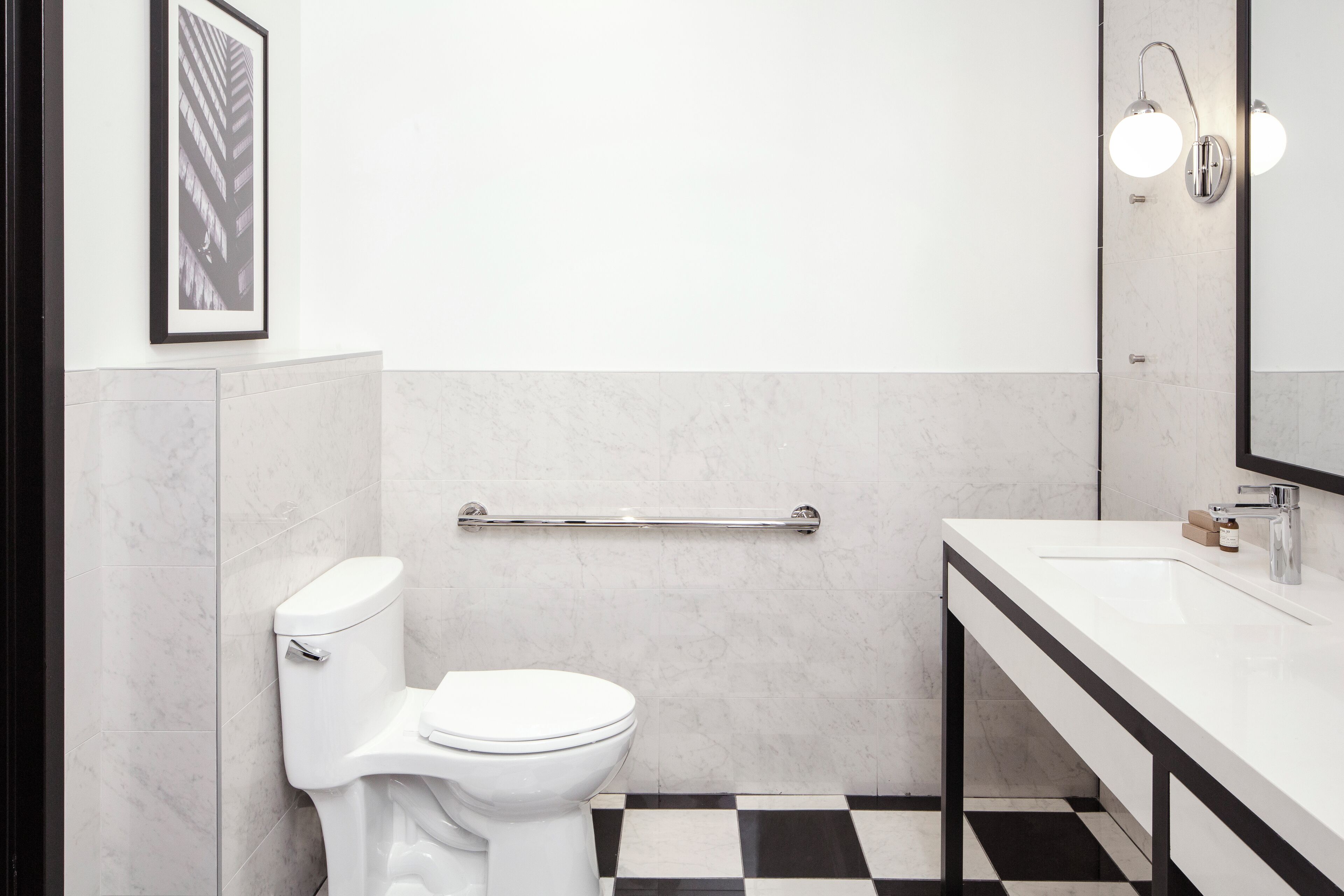 Un baño moderno con inodoro blanco, suelo a cuadros blanco y negro, lavamanos blanco con espejo arriba y un cuadro enmarcado en la pared.