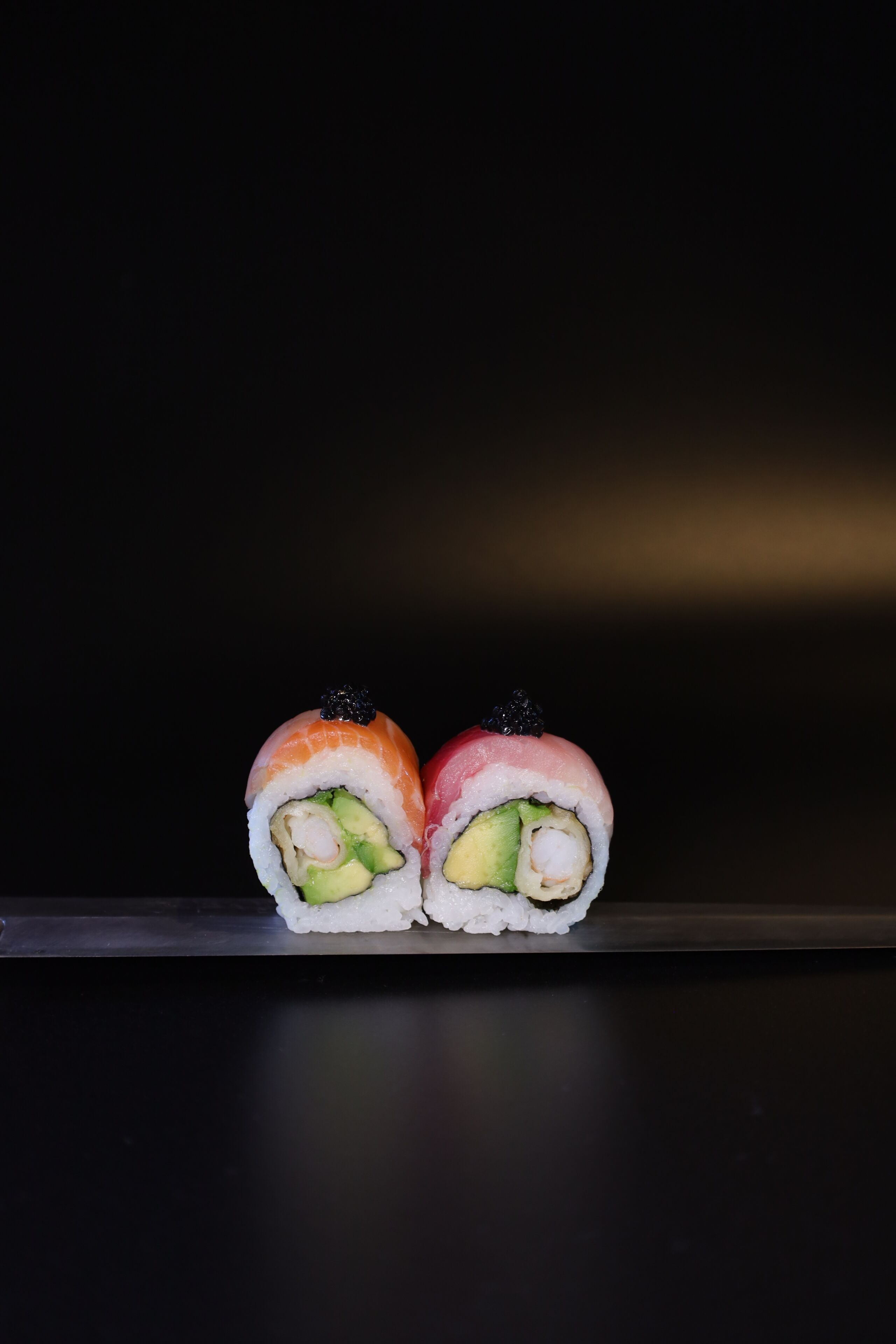 Two sushi rolls with avocado and salmon, placed on a dark surface.
