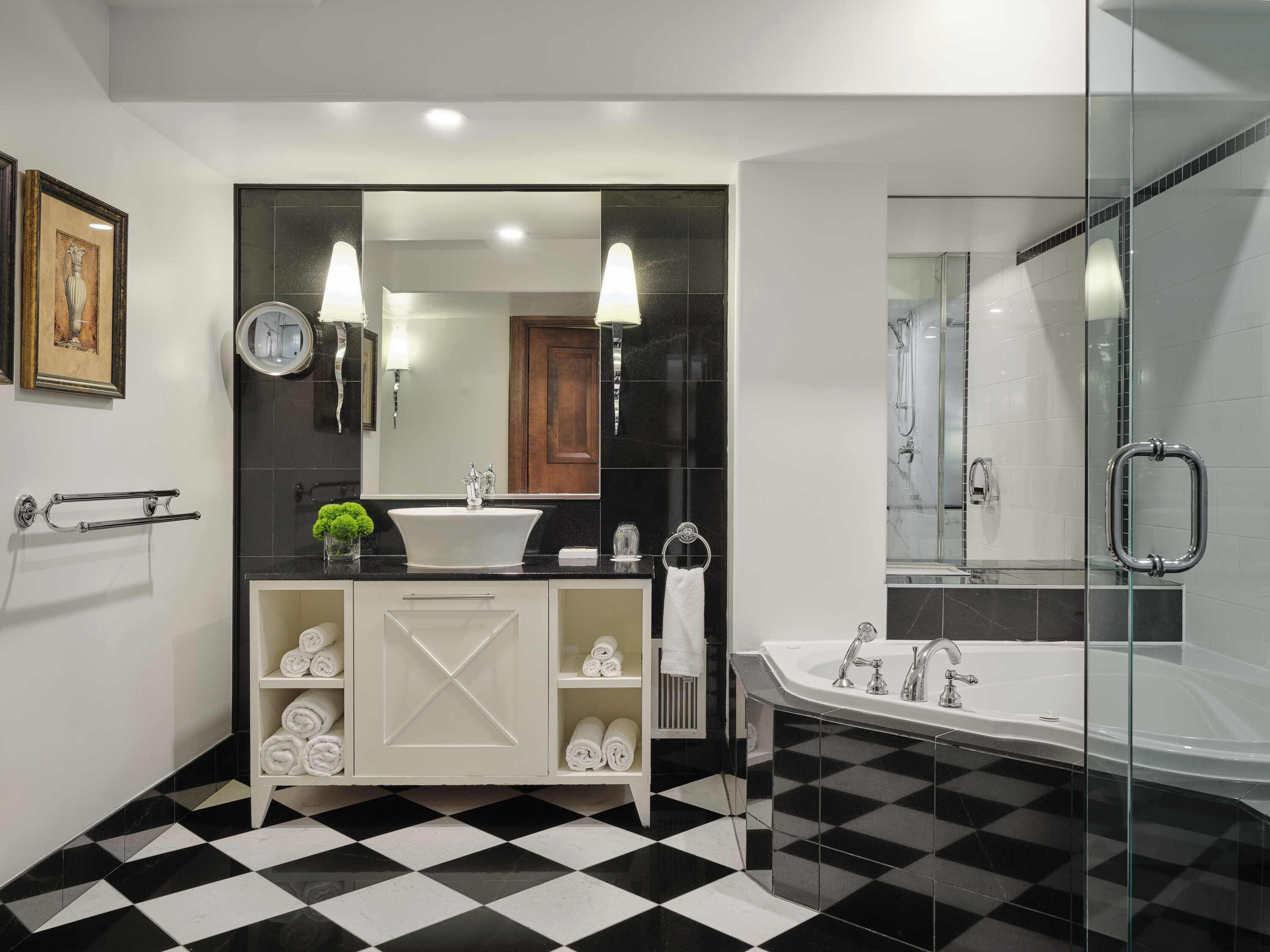 Un baño moderno con piso a cuadros blanco y negro, un tocador blanco con espejo y zona de ducha con mampara de vidrio.