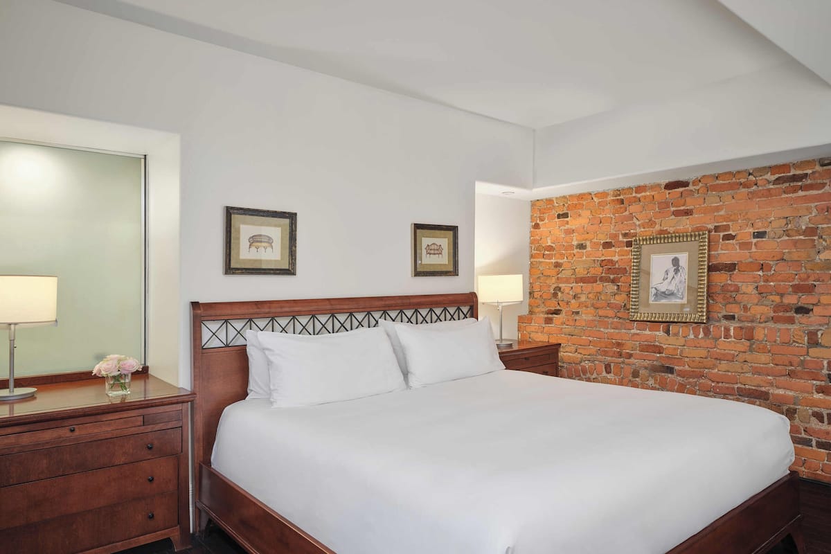 A neatly made bed with white linens, a wooden headboard, and a brick wall behind it. There are two framed pictures on the wall, a nightstand with a lamp, and a dresser with drawers.