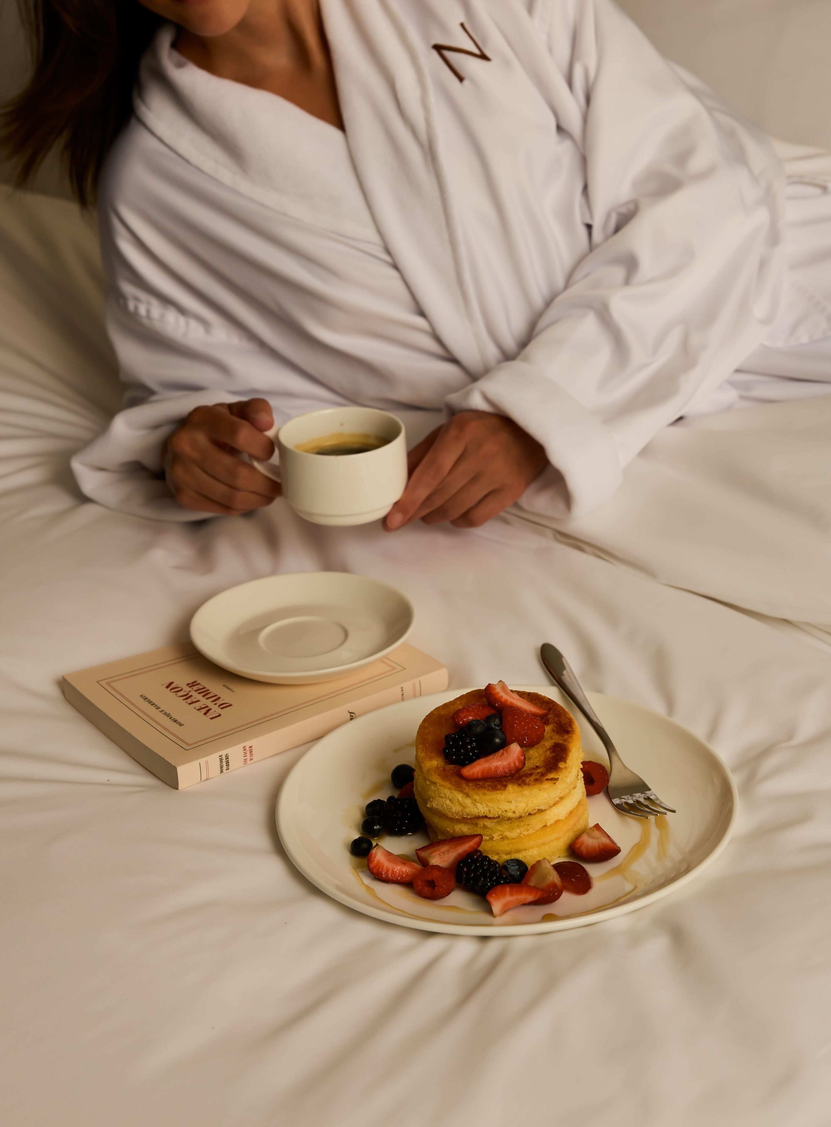 En person i en vit dräkt som håller i en kopp, en tallrik med pannkakor och bär samt en bok som ligger på en säng.