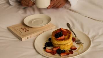 A person in a white robe holding a cup, a plate with pancakes and berries, and a book on a bed.