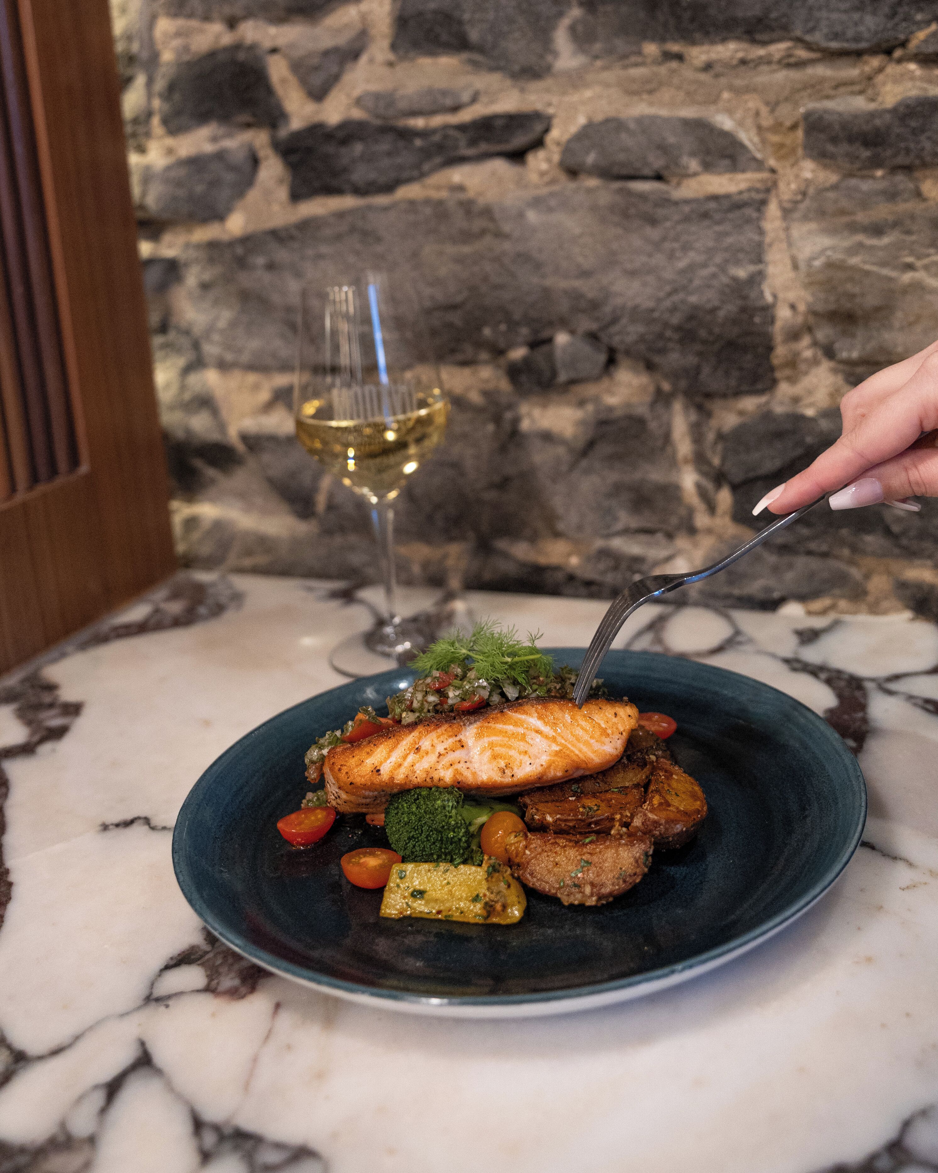 A plate of food with a fork, a glass of white wine, and a stone wall background.