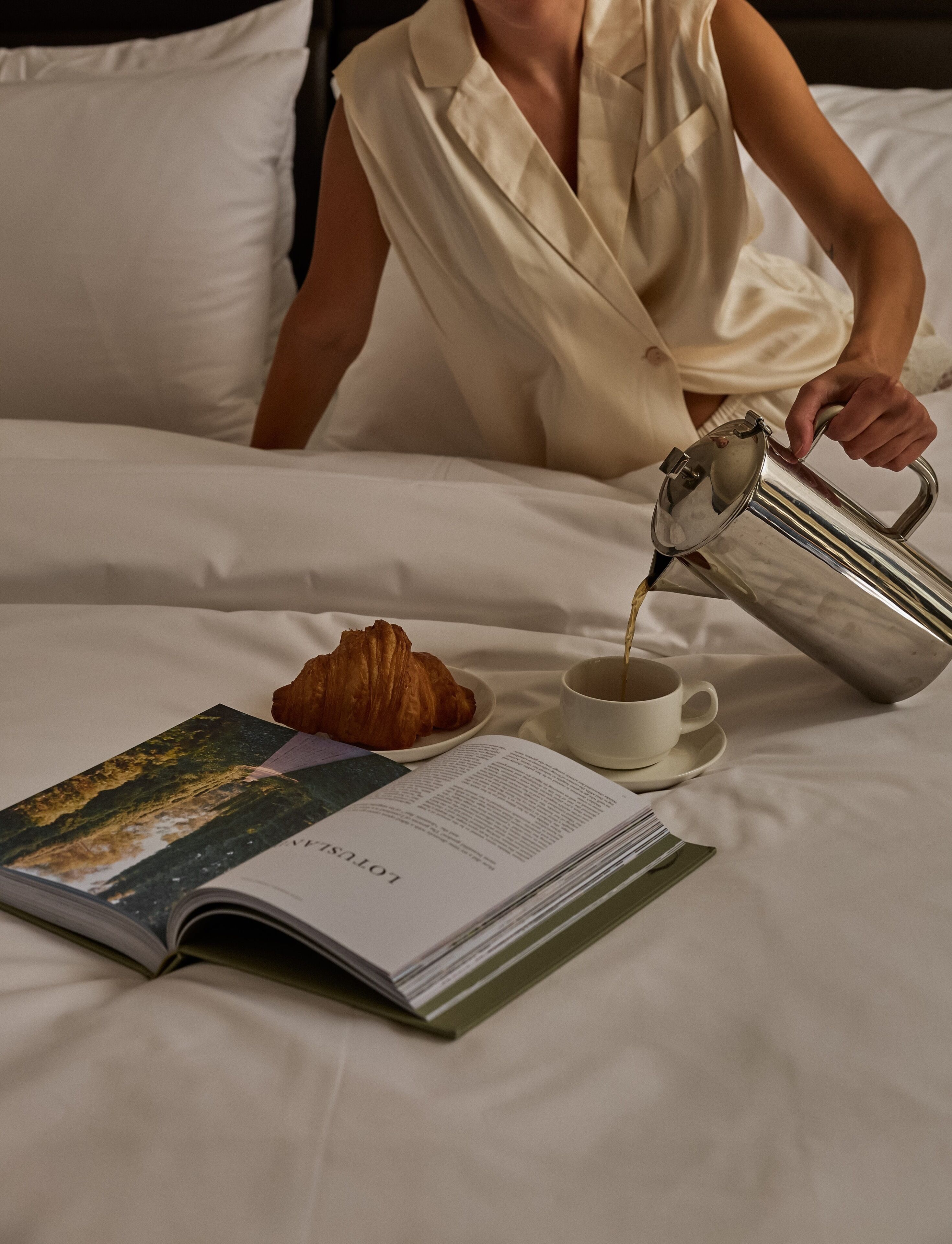 A person pouring coffee into a cup on a bed with a book and a croissant.