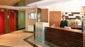 Zona de recepción de un hotel con un mostrador de madera, una recepcionista, una planta y una puerta roja al fondo.
