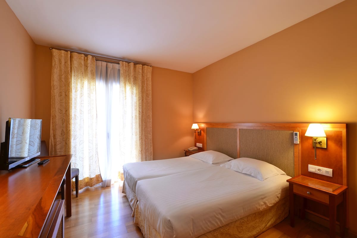 A hotel room with two beds, a desk with a television, and a bedside table with a lamp.