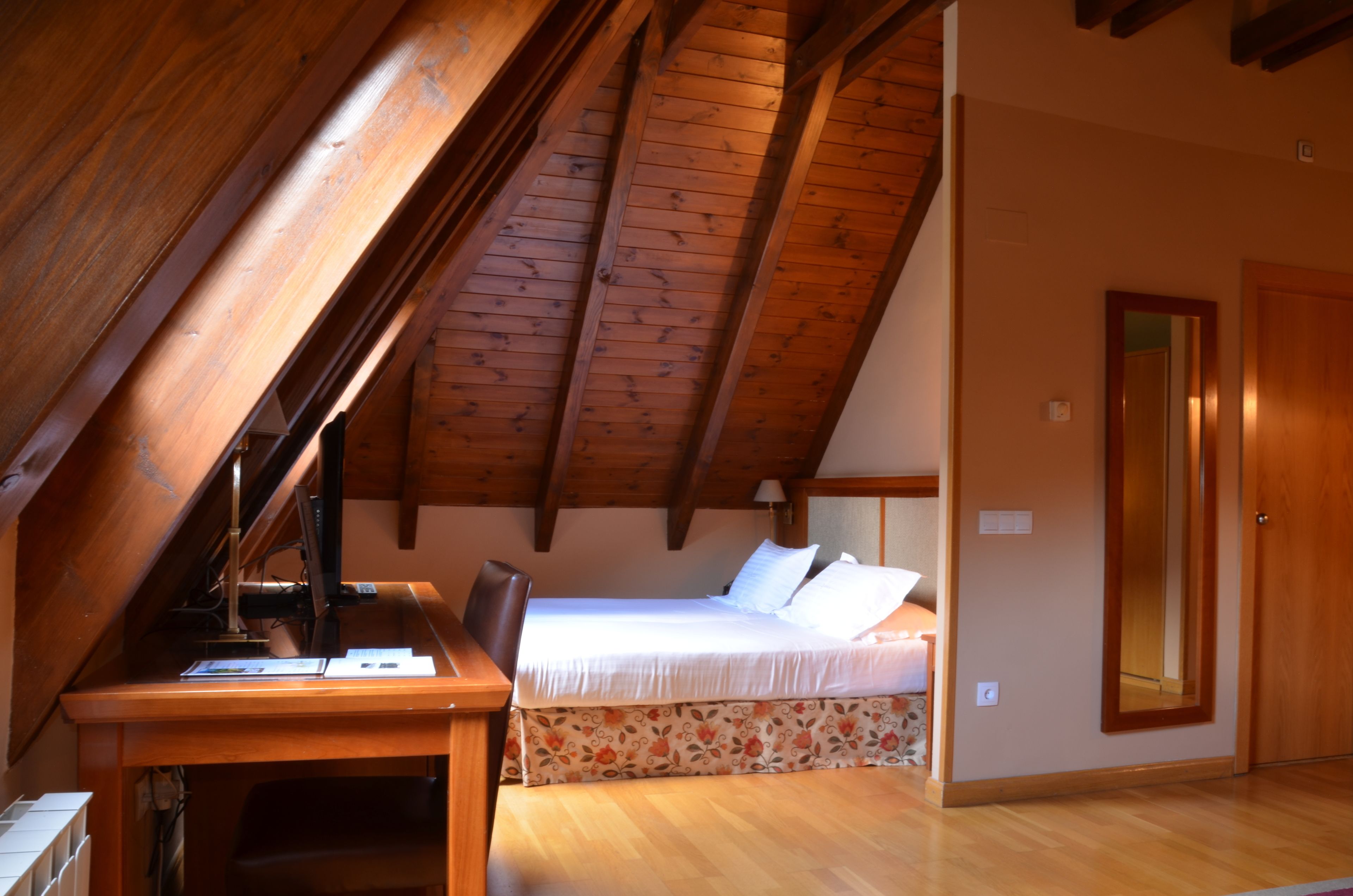 Un dormitorio con techo inclinado, vigas de madera, una cama con ropa blanca, un escritorio con silla y un espejo en la pared.