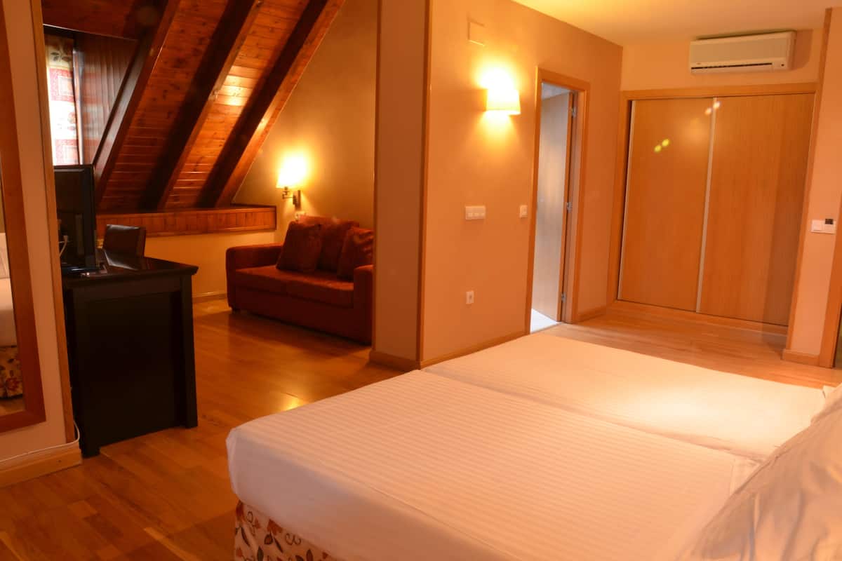 A bedroom with a large bed, a desk, a chair, and a sofa. There is a wardrobe and an air conditioning unit on the wall.