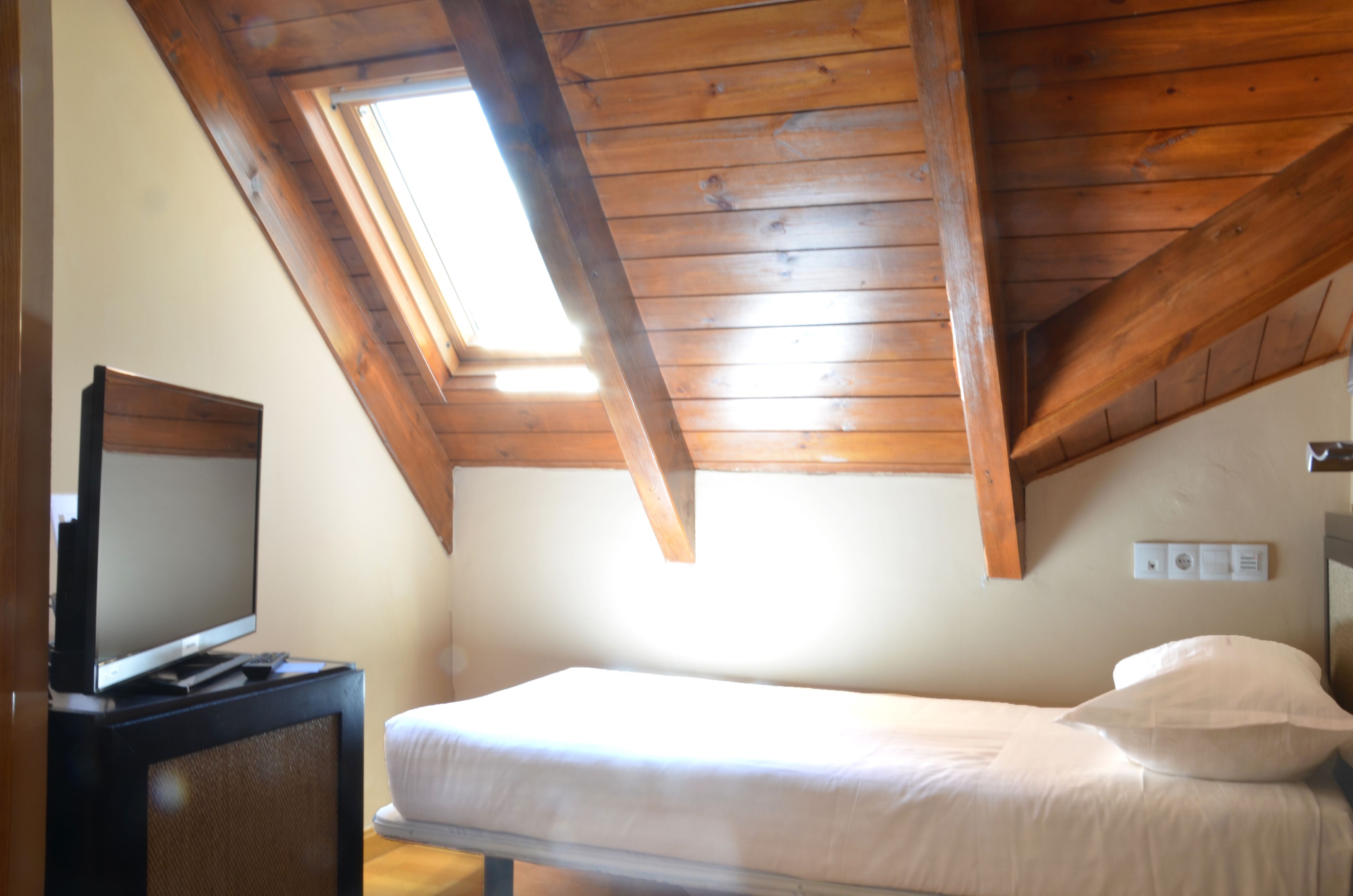 Een kamer met een schuin houten plafond, een bed met wit beddengoed, een televisie op een standaard en een raam voor natuurlijk licht.