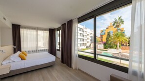 Una habitación de hotel con cama, un pequeño sofá y ventana con vista a una piscina y edificios.
