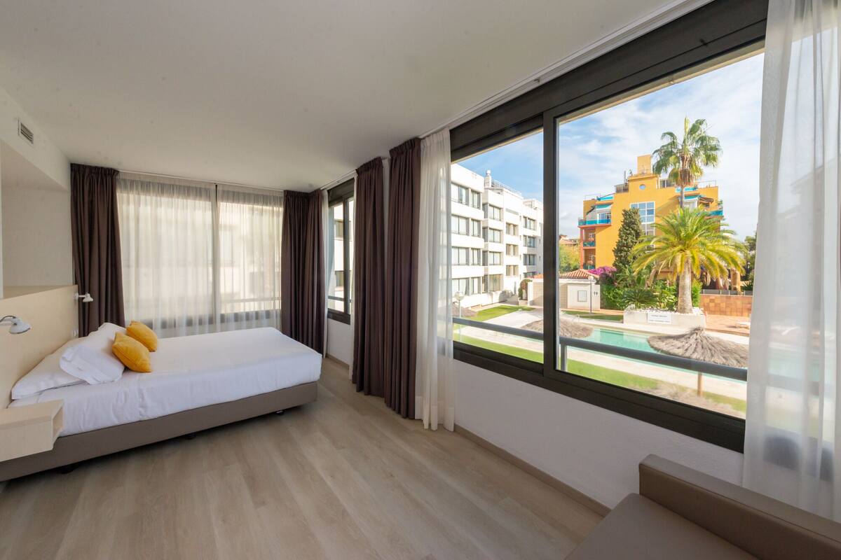 Una habitación de hotel con cama, un pequeño sofá y ventana con vista a una piscina y edificios.
