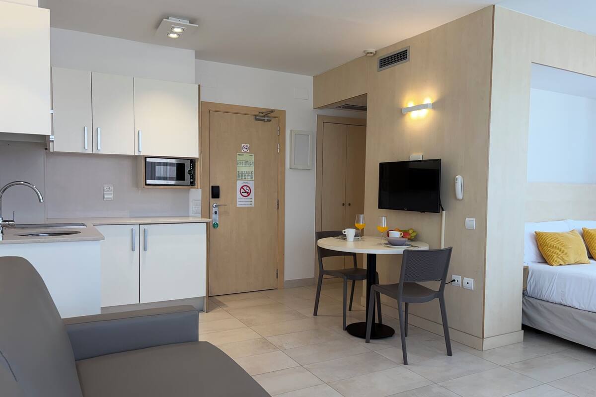 Una habitación de hotel moderna con kitchenette, zona de comedor, televisión y cama con almohadas amarillas.