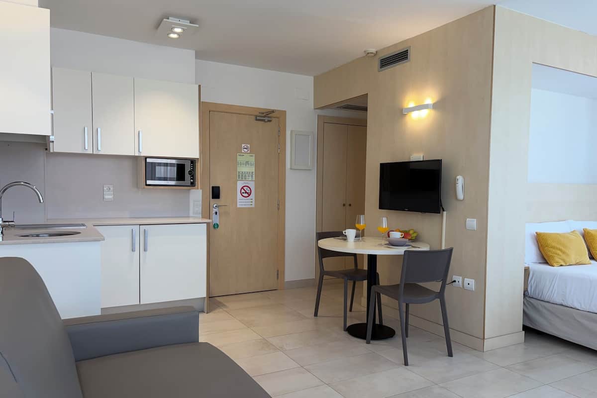 A compact hotel room with a kitchenette, a dining area, a flat-screen TV, and a bed with yellow pillows.