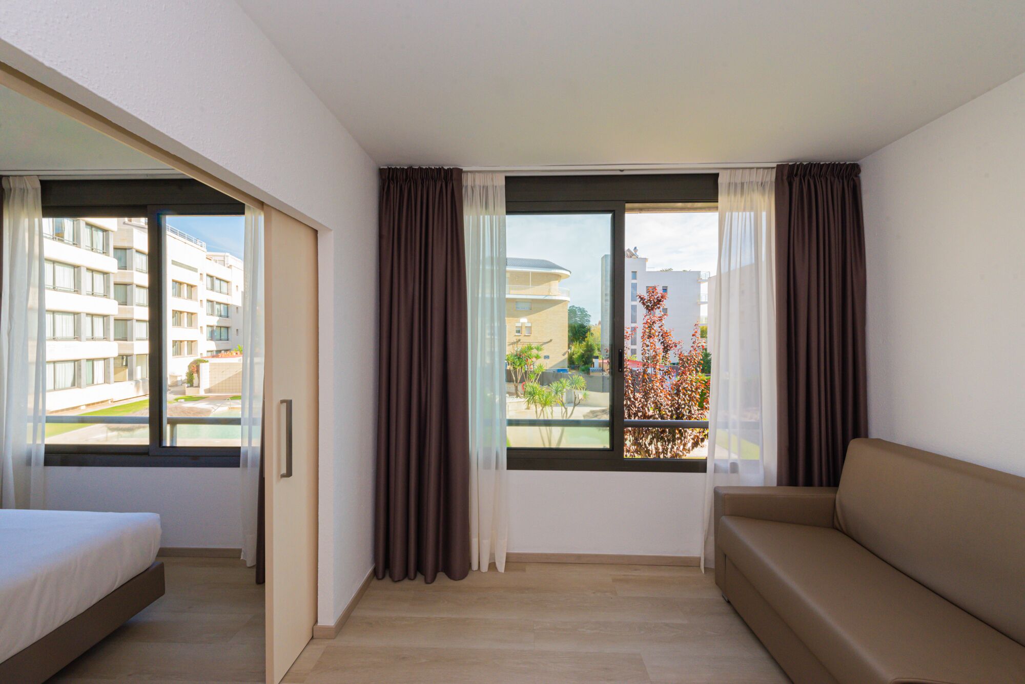 Quarto de hotel moderno com cama, sofá e janelas amplas com vista para a cidade.