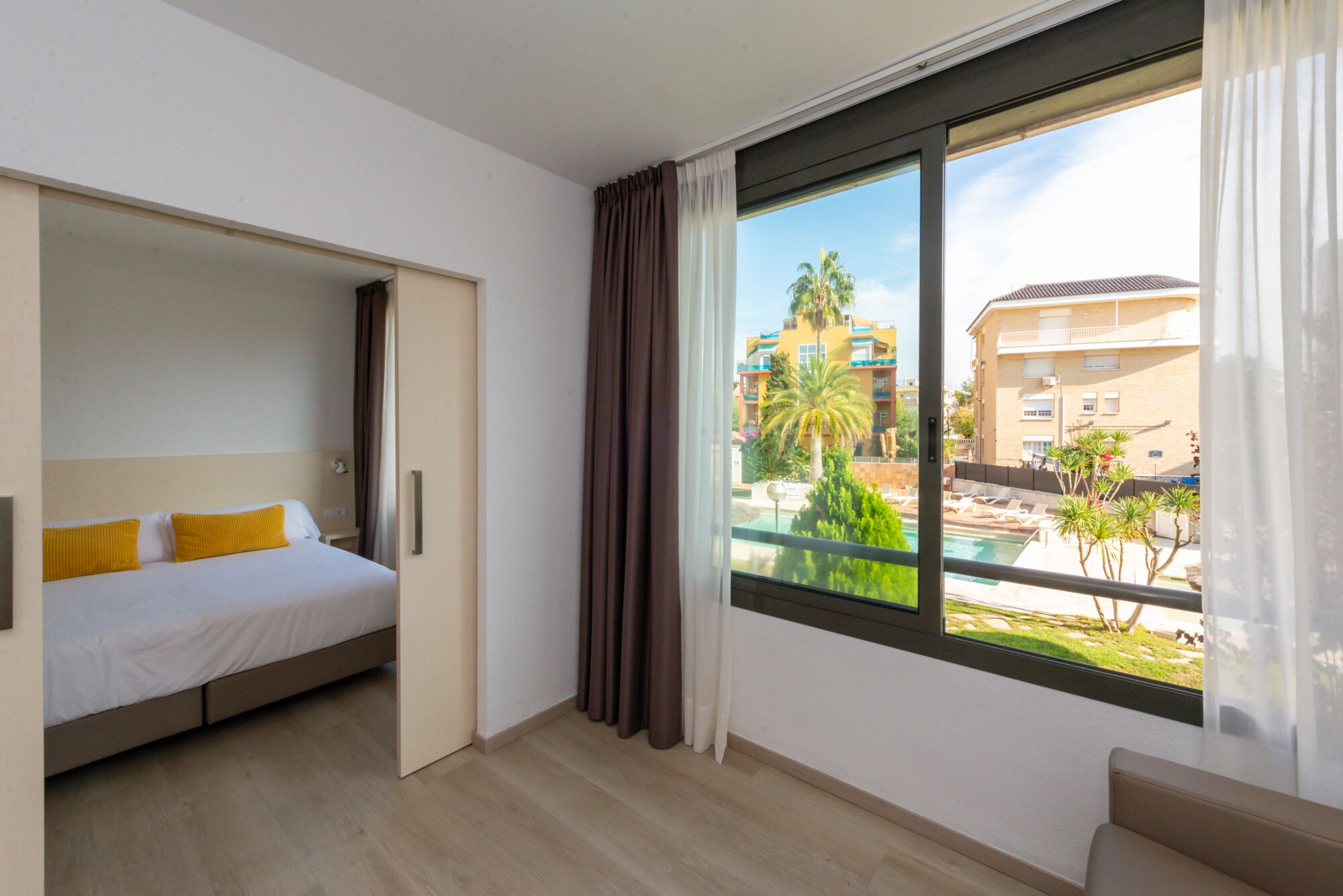 Quarto de hotel com cama, um pequeno espaço de estar e uma janela grande com vista para uma piscina e edifícios.