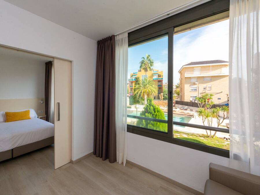Quarto de hotel com cama, um pequeno espaço de estar e uma janela grande com vista para uma piscina e edifícios.