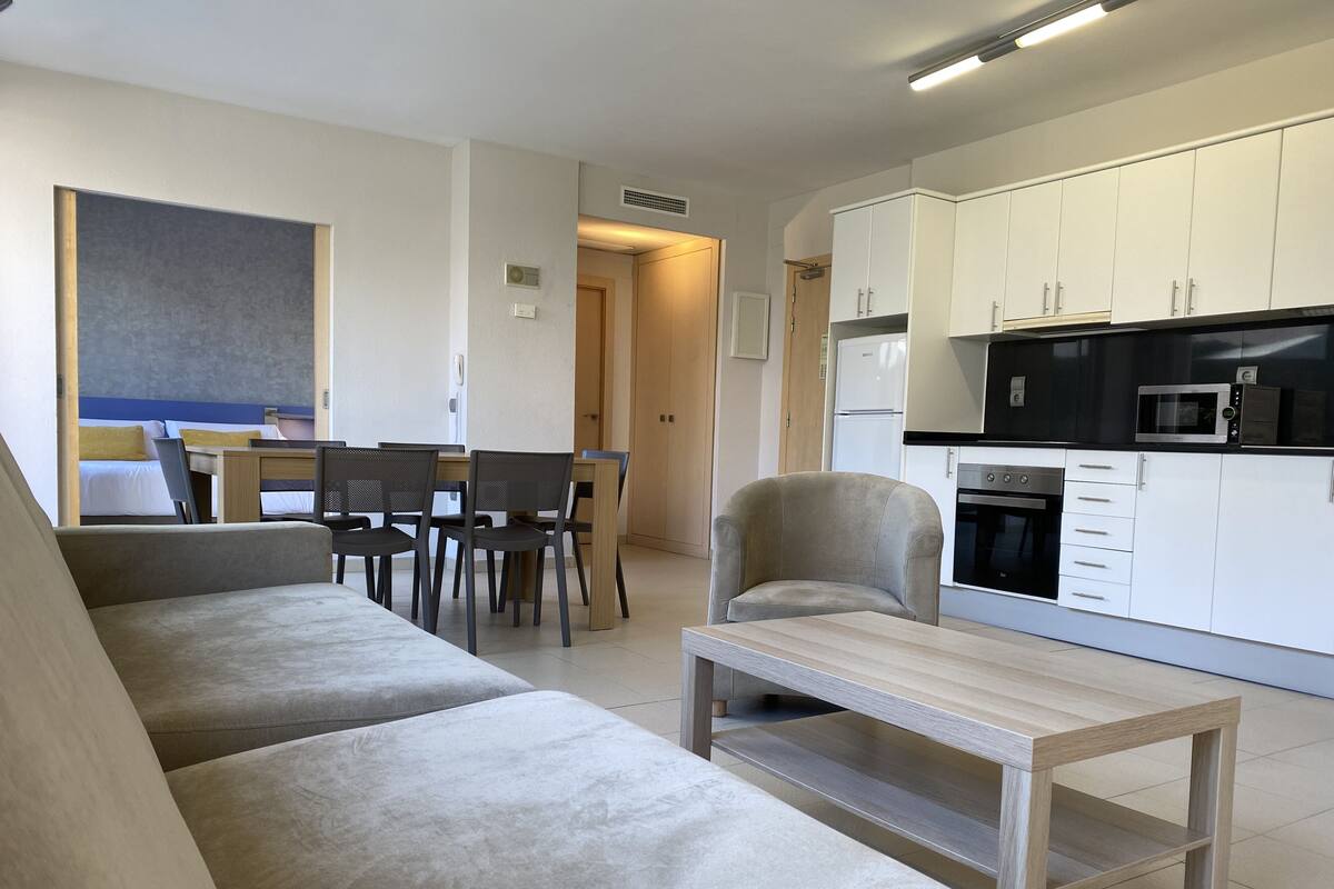 Una sala de estar moderna con sofá, mesa de comedor, sillas y una cocina con armarios y electrodomésticos blancos.