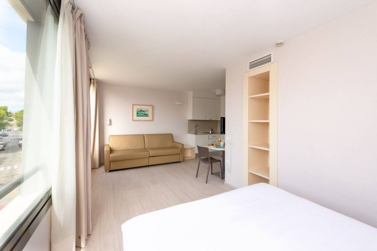 Habitación de hotel con cama, sofá, escritorio con silla, kitchenette y ventana con cortinas.