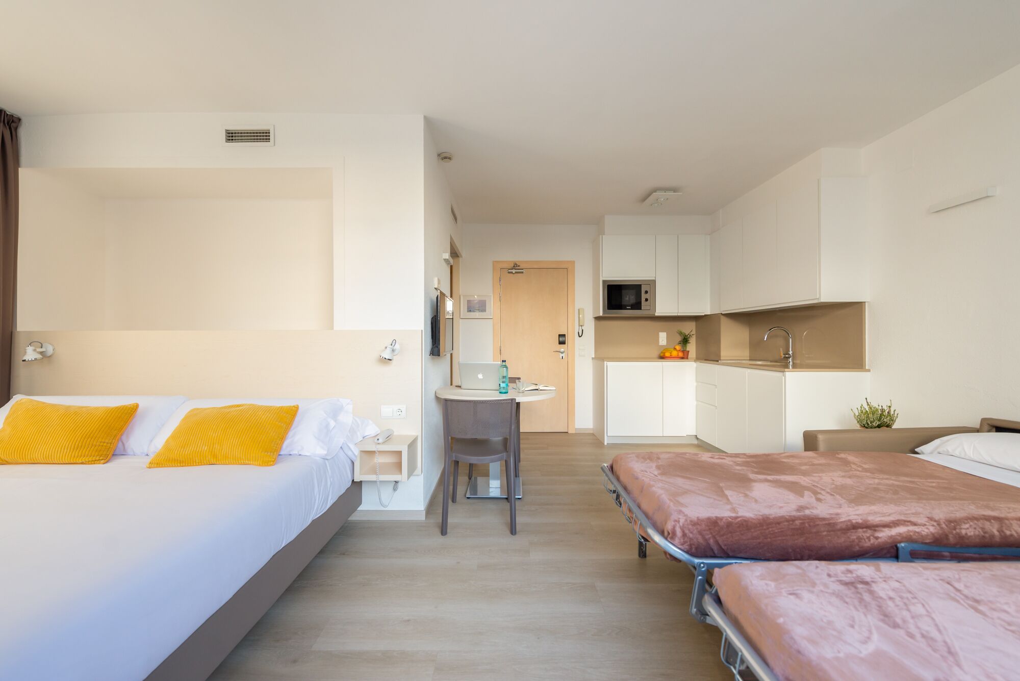 A hotel room with two beds, a desk with a laptop, a microwave, and a small kitchenette.