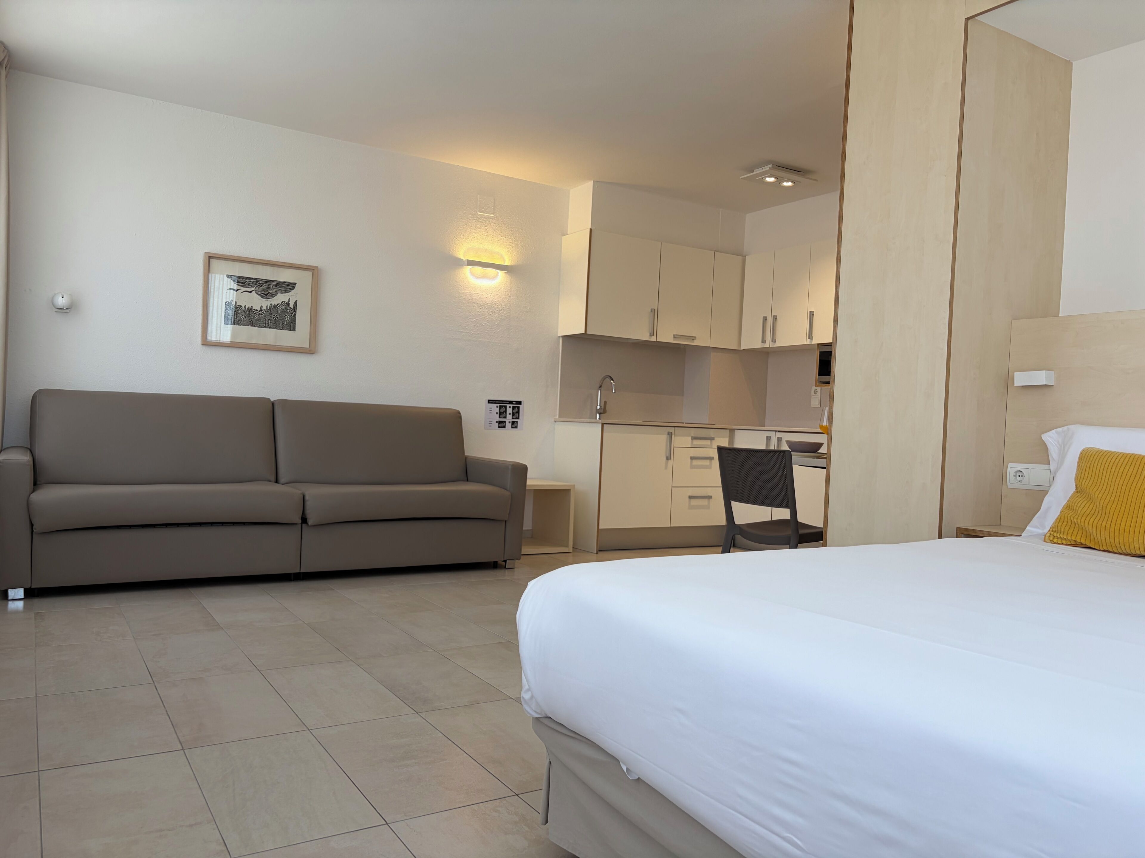 A hotel room with a bed, a sofa, a chair, a kitchenette, and a wall-mounted light fixture.