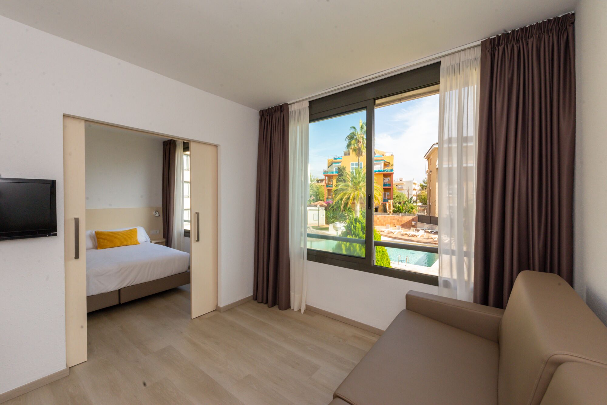 Ein Hotelzimmer mit Bett, Fernseher, Sofa und großem Fenster mit Blick auf einen Pool und Gebäude.