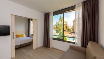 Ein Hotelzimmer mit Bett, Fernseher, Sofa und großem Fenster mit Blick auf einen Pool und Gebäude.