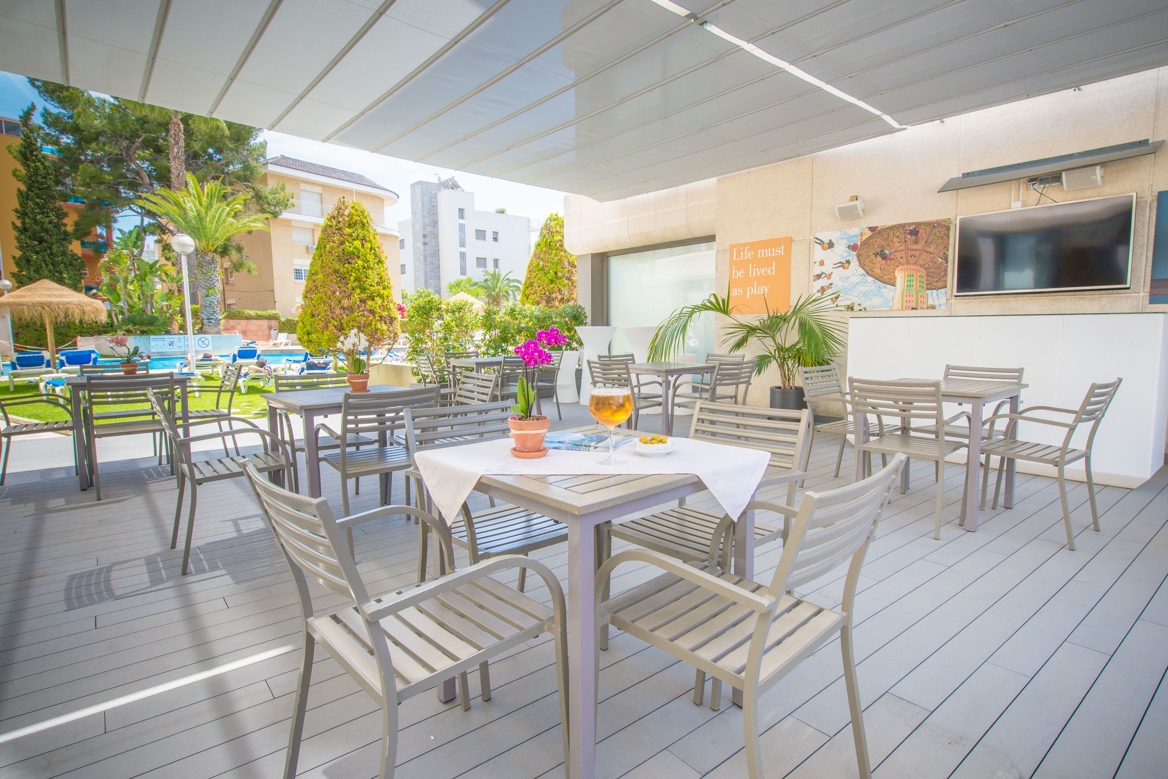 Un área de comedor al aire libre con mesas y sillas, rodeada de plantas en macetas y una piscina.