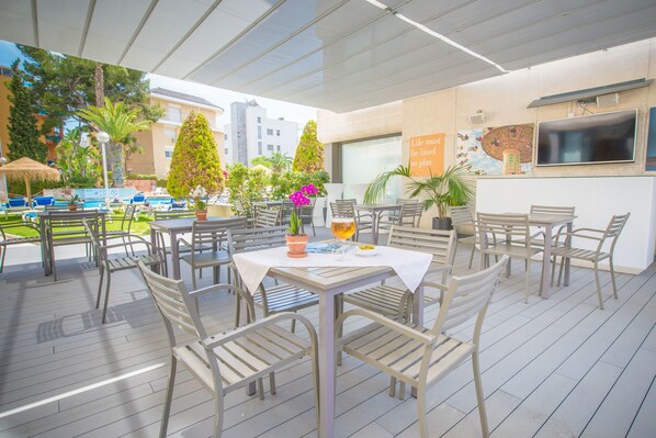 Un espace extérieur aménagé pour les repas, avec des tables et des chaises, entouré de plantes en pot et d’une piscine.