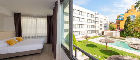 A hotel room with a bed, a view of a swimming pool, and a building with balconies.