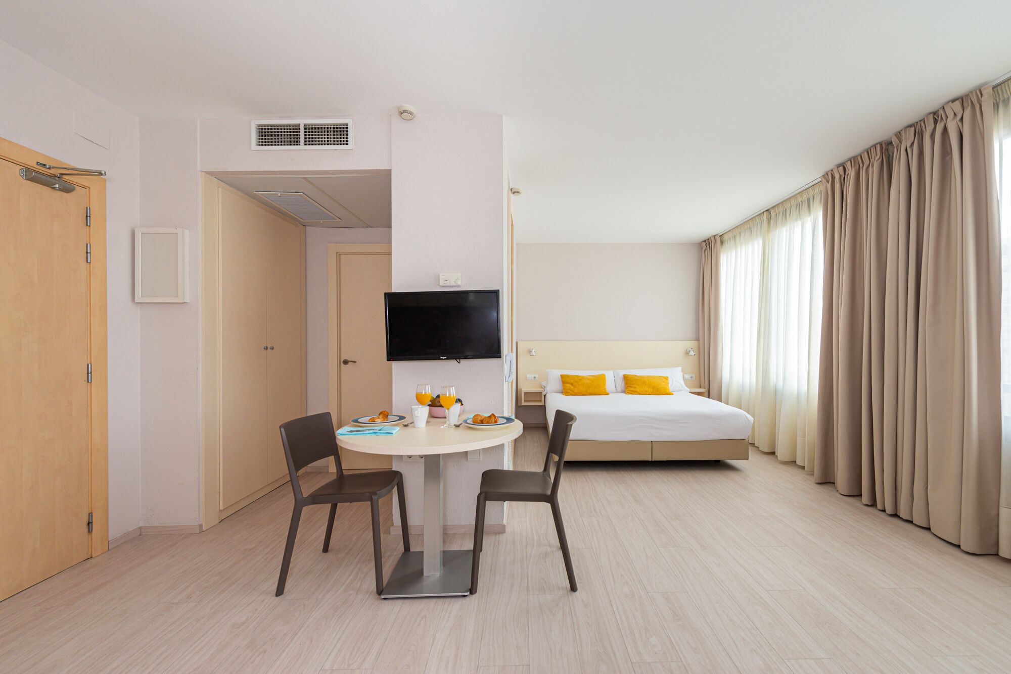 Une chambre d’hôtel comprenant un lit, une télévision, une table à manger avec des chaises et une fenêtre avec des rideaux.