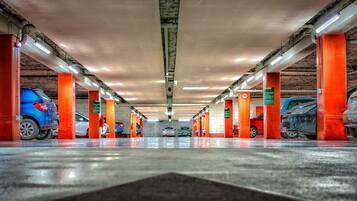 Un aparcamiento de varios niveles con coches estacionados en espacios designados, iluminado por luces superiores y sostenido por pilares anaranjados.