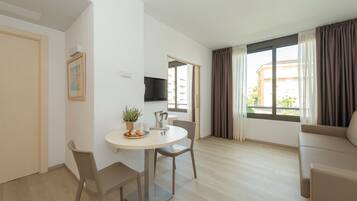 Una sala de estar moderna con una mesa redonda de comedor, dos sillas, un sofá, un televisor montado en la pared y una ventana con cortinas.