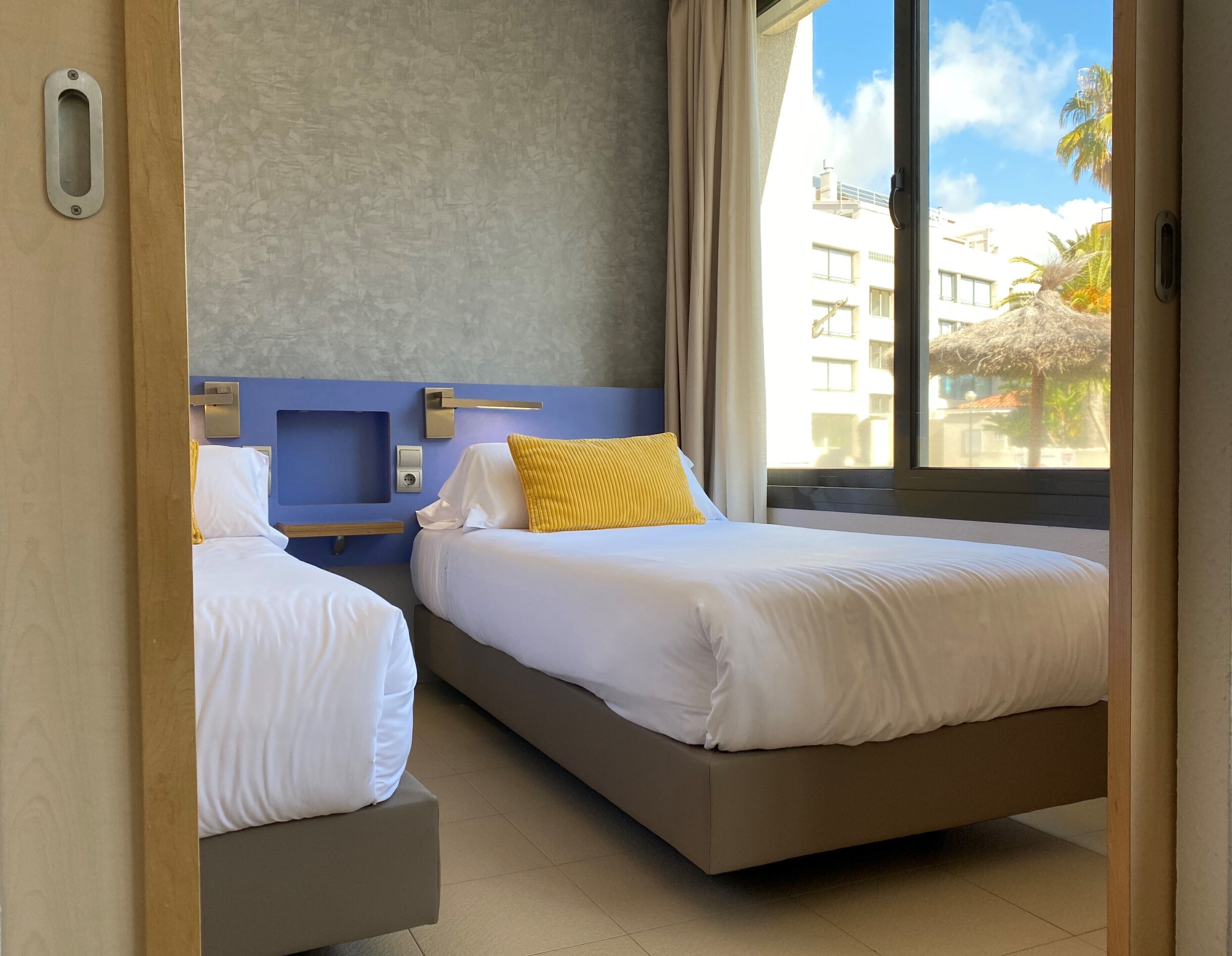 Een hotelkamer met twee bedden, een blauwe accentmuur en een groot raam met uitzicht op gebouwen en palmbomen.