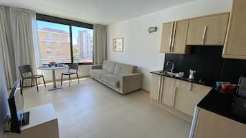 Een ruimtelijke leefruimte met kitchenette, een eethoek met tafel en stoelen, een bank en een groot raam met uitzicht op gebouwen.