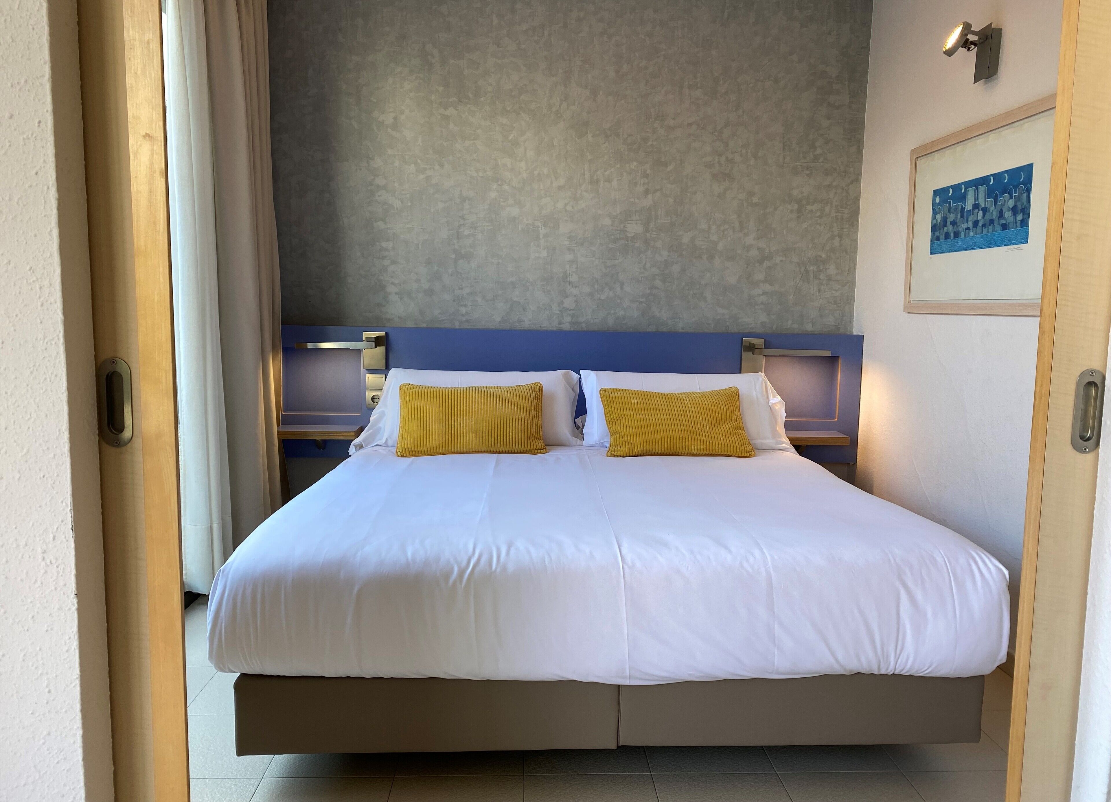A double bed with white bedding and two yellow pillows, a nightstand with a lamp, and a framed picture on the wall.