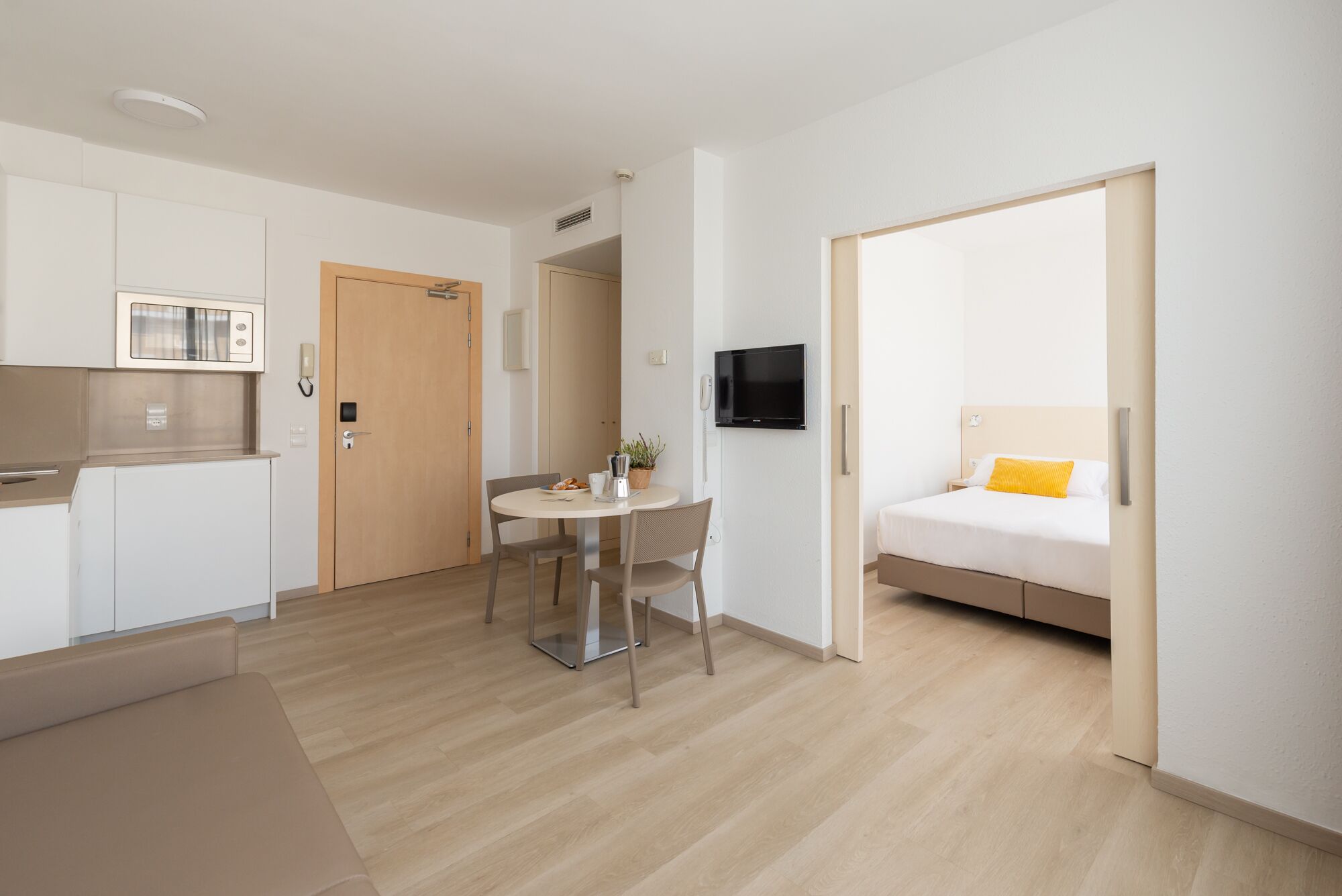 A modern hotel room with a bed, a small dining table, a kitchenette, and a TV.