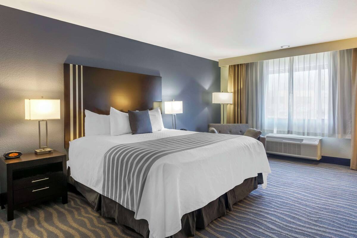 A hotel room with a large bed, two bedside tables with lamps, a chair, a window with curtains, and an air conditioning unit.