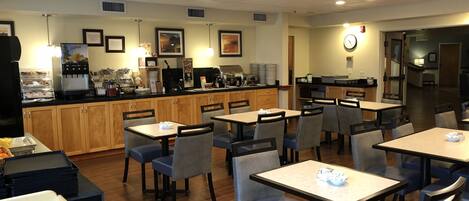 A clean, well-lit dining area with multiple tables and chairs, a coffee station, and framed pictures on the walls.