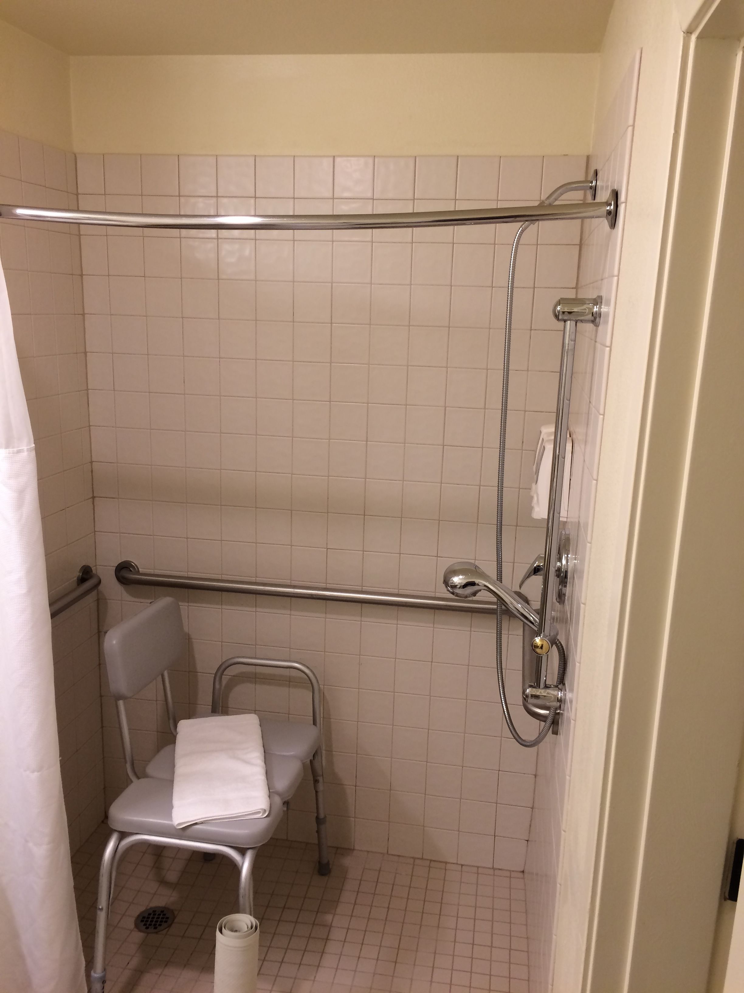 Un pequeño espacio de ducha con azulejos, una barra de apoyo, un cabezal de ducha y una silla con una toalla.
