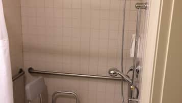 Un pequeño espacio de ducha con azulejos, una barra de apoyo, un cabezal de ducha y una silla con una toalla.