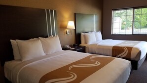 A hotel room with two beds, each with white linens and a brown decorative stripe. There is a nightstand with a lamp and a phone between the beds. A large window provides a view of greenery outside. - Quality Inn & Suites Bainbridge Island (Bainbridge Island)
