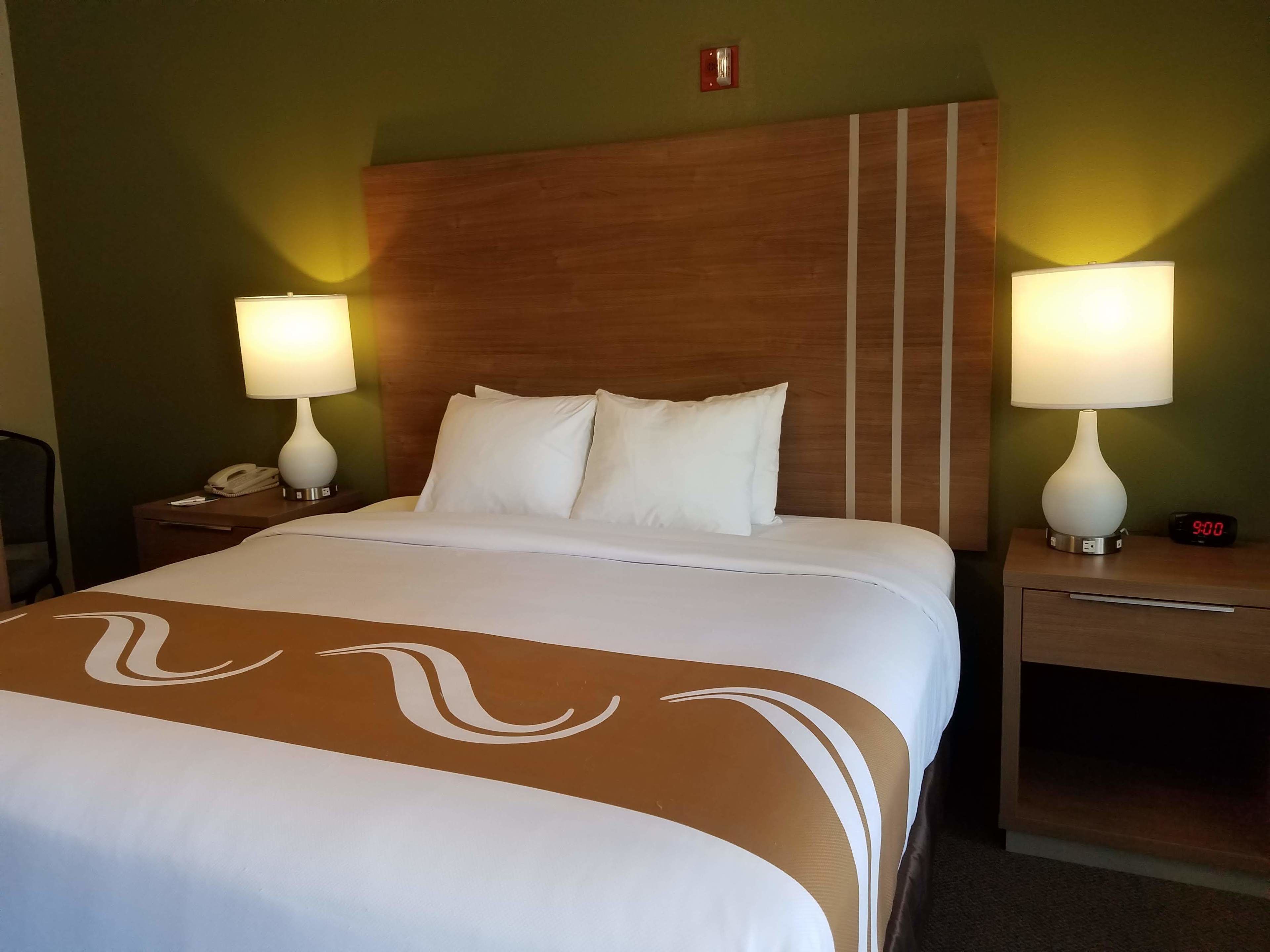 A hotel room with a large bed, two bedside tables with lamps, a nightstand, and a wall-mounted alarm clock.