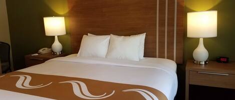 A hotel room with a large bed, two bedside tables with lamps, a nightstand, and a wall-mounted alarm clock.