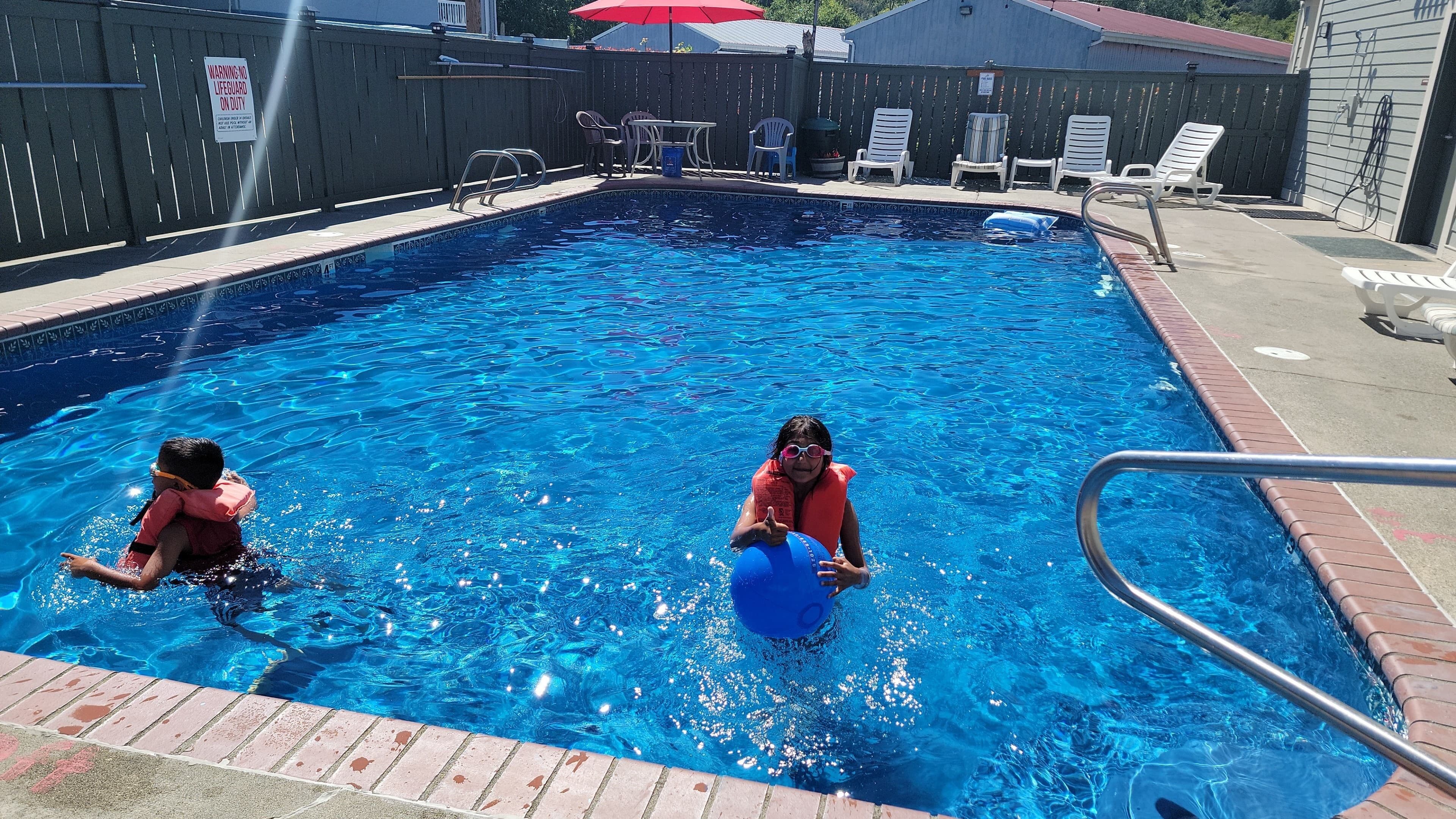 A swimming pool with two children playing, surrounded by lounge chairs and a fence.